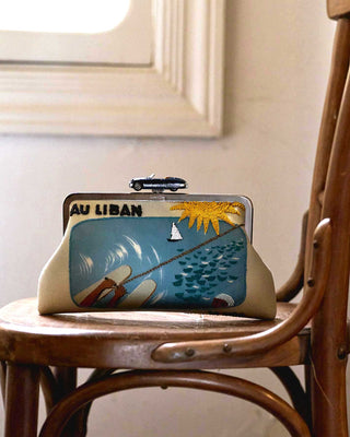 A vintage-style clutch bag with a colorful travel-poster design reading 'AU LIBAN' sits on a wooden chair in front of a window.