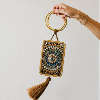 Hand holding a decorative handbag with intricate design and tassel on a plain background
