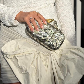 A close-up of a person holding a metallic silver textured clutch bag with a gold accent, wearing a white textured sweater and a cream pleated skirt.