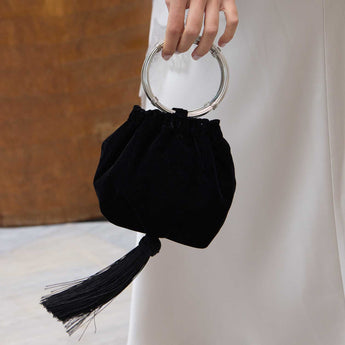 Black round  handbag with a ring handle and a tassel held by a hand against a neutral background.