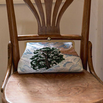 A vintage-style clutch purse with a gold-tone metal frame and clasp, featuring a printed design of a snowy pine tree on a swirling blue and white sky background. The purse is displayed on a wooden chair.
