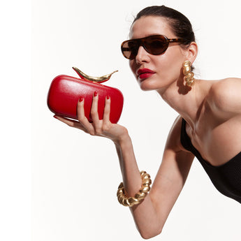 A red clutch bag handed by a model with a sleek finish and a gold-dipped clasp shaped like a chili pepper.