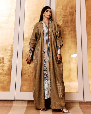 A person wearing an olive-colored abaya with hand-crocheted details on the front lapels and sleeve slits, standing in front of a golden background.