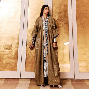 A person wearing an olive-colored abaya with hand-crocheted details on the front lapels and sleeve slits, standing in front of a golden background.