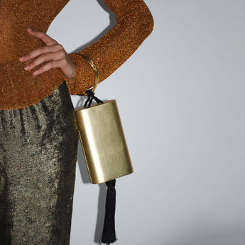 Gold handbag with tassel held by a person wearing a brown textured sweater and green skirt on a light gray background