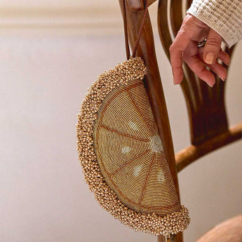 A close-up of a hand holding a semi-circular beaded handbag with a brown leather strap, hanging from a wooden chair in an indoor setting.