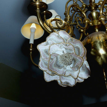A wristlet handbag designed in the shape of a blooming magnolia flower, with gold, pink, and white colors and a gold-tone metal handle hanging from a chandelier