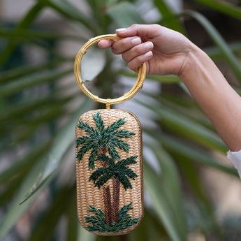 A hand holding straw gold straw bag with a palm tree motif and green glass beads, featuring twin gold bracelet handles.