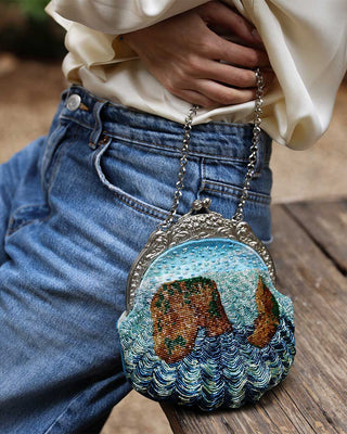 A close-up lifestyle shot of a person wearing blue jeans and a cream blouse, holding a small round embroidered clutch purse with a silver-tone chain strap. The purse features a textured design resembling waves and a brown landscape under a light blue sky.