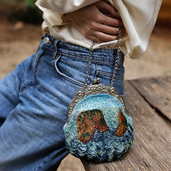 A close-up lifestyle shot of a person wearing blue jeans and a cream blouse, holding a small round embroidered clutch purse with a silver-tone chain strap. The purse features a textured design resembling waves and a brown landscape under a light blue sky.