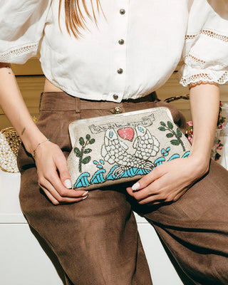 Person holding a handbag with embroidered design of two fishes holding a heart in a casual setting.