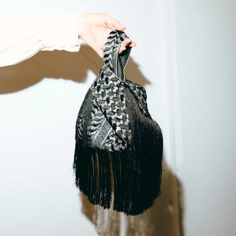 Hand holding a black and gray keffiyeh patterned scarf with fringe on a white background