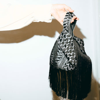 Hand holding a black and gray keffiyeh patterned scarf with fringe on a white background