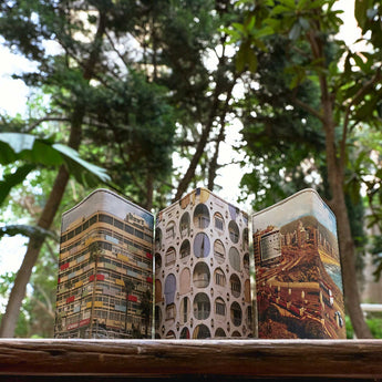 Three small passport cover printed with architectural building designs, arranged side-by-side on a wooden surface with a blurred background of trees.
