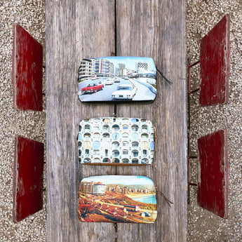 Three zippered coin pouches with printed scenic designs are arranged vertically on a weathered wooden plank outdoors, flanked by red wooden panels and gravel.