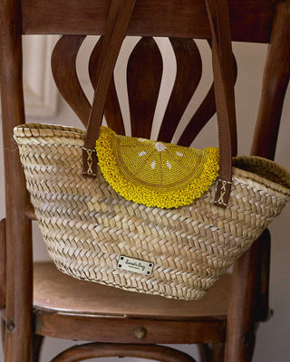 A woven straw tote bag with brown leather handles and a bright yellow beaded semicircle accent on the front, resting on a dark wooden chair.