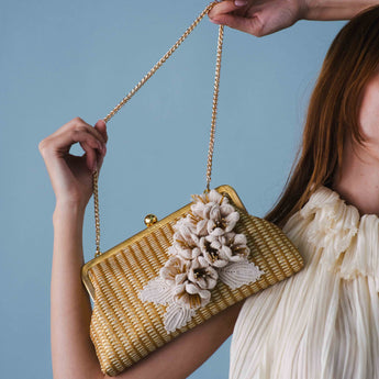Woman holding a gold clutch with crocheted floral embellishments on straw on a blue background