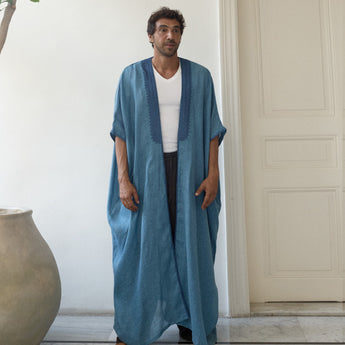 Man wearing a long blue abaya over a white shirt in a minimalistic room.