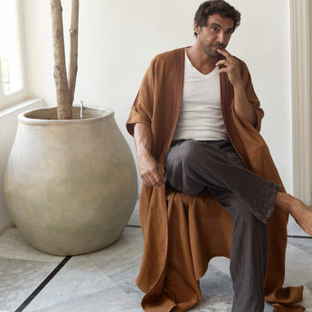 Man sitting on a stool wearing a brown abaya in a room with a large plant.
