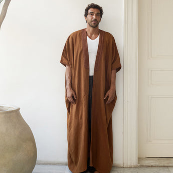Man wearing a brown abaya over a white shirt in a minimalistic setting.