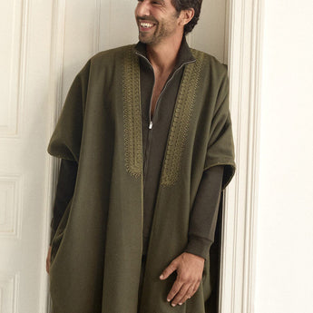 Man wearing a long green abaya standing against a white wall.