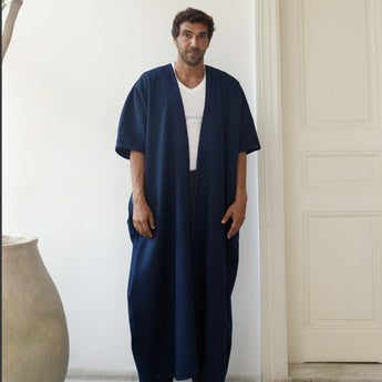 Man wearing a navy blue abaya over a white shirt in a minimalistic room.