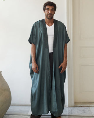 Man wearing a long teal abaya over a white shirt in a minimalistic room.