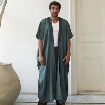 Man wearing a long teal abaya over a white shirt in a minimalistic room.