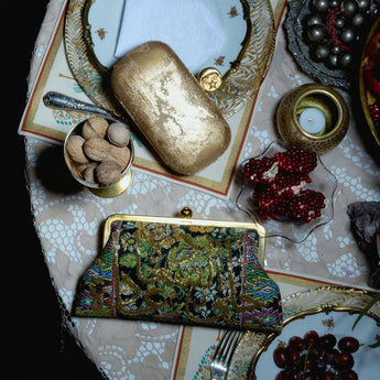 Decorative table setting with gold clutch, patterned purse, and fruit on a textured surface.