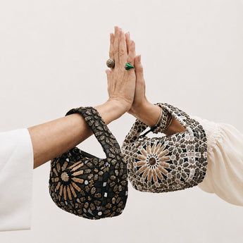 Two hands with intricately Moroccan designed bangles and rings, wearing patterned sleeves against a neutral background.