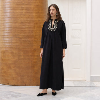 Woman wearing a black cotton amira dress with a decorative  beaded petal neckline in a minimalistic setting.