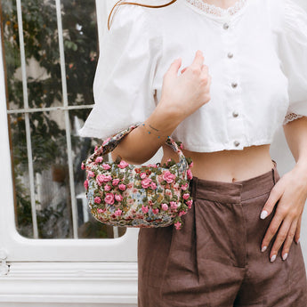 Person holding a floral patterned with crocheted pink roses handbag with a blurred outdoor background.