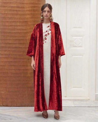 Woman wearing a red velvet robe over a white vanda with floral patterns, standing against a wooden wall.