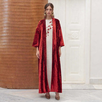 Woman wearing a red velvet robe over a white vanda with floral patterns, standing against a wooden wall.