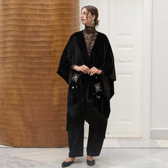 Woman wearing a black velvet poncho with embroidered sunrise details in a minimalistic setting.