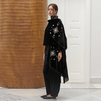 Woman wearing a black velvet poncho with embroidered sunrise details and a fringed shawl in an indoor setting.