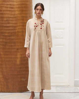 Woman wearing a long, beige amira with floral embroidery in a minimalistic setting.
