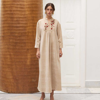 Woman wearing a long, beige amira with floral embroidery in a minimalistic setting.