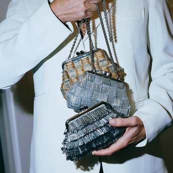 Person holding two woven handbags, one silver and one with a patterned strap, against a neutral background.