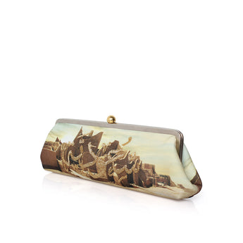 A rectangular clutch purse with a gold-tone metal frame and clasp, featuring a printed design of a castle on a beach with gold glittery waves. The clutch is photographed on a clean white background with a soft reflection underneath.