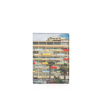 A slim passport cover with a printed photograph of a mid-century modern apartment building, featuring colorful balconies and palm trees, shown on a plain white background.