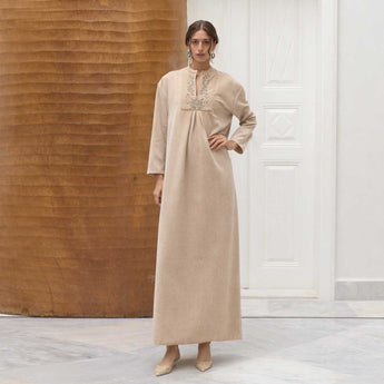 Woman wearing a beige long Amira dress with intricate detailing in a minimalistic setting.