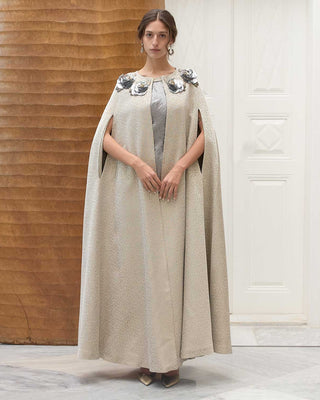 Woman wearing a long, light-colored cape with flower elements in a minimalistic setting.