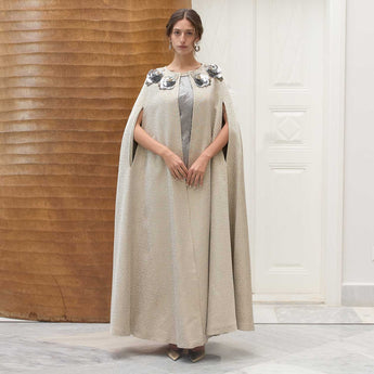 Woman wearing a long, light-colored cape with flower elements in a minimalistic setting.
