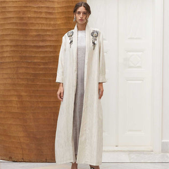 Woman wearing a long, light-colored cover up with floral embellishments in a minimalistic setting.