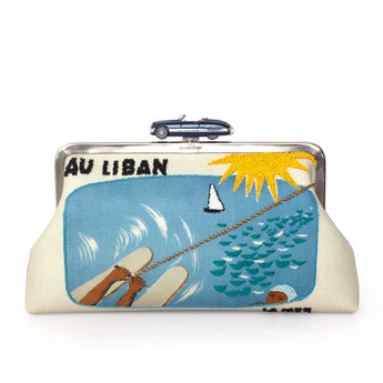 An embroidered clutch purse with a silver metal frame and a blue convertible clasp. The front features a colorful embroidered design depicting a beach scene with the text 'AU LIBAN' and a small car charm resting on top.