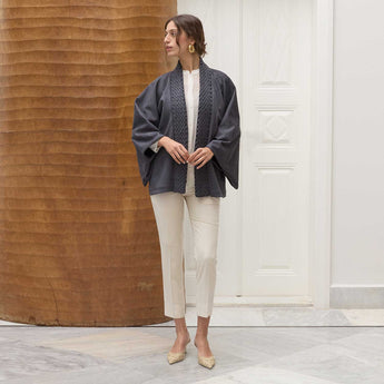 Woman wearing a navy blue jacket over a white top and beige pants, standing against a wooden wall.