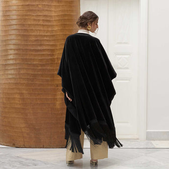 Woman wearing a black fringed poncho over a white shirt and beige pants in a minimalistic setting.