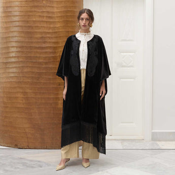 Woman wearing a black fringed poncho over a white shirt and beige pants in a minimalistic setting.