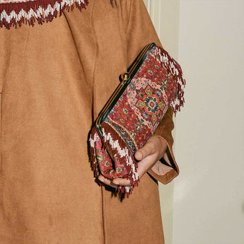 Model in a camel outfit holding the Tapestry Red Le Long clutch, showing its ornate pattern, metal clasp and red-and-white beaded fringe.